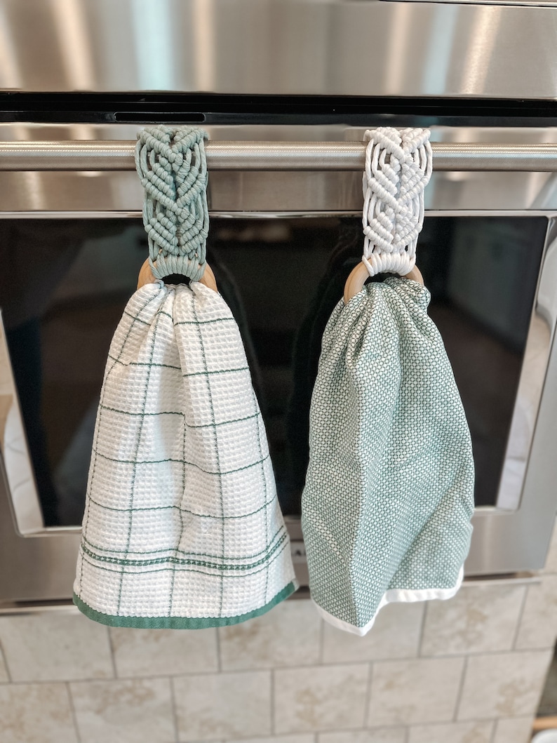 May include: Two green kitchen towels hanging on a silver oven handle. The towels are held in place by macrame loops with wooden rings. One towel has a white and green checkered pattern, and the other has a solid green pattern.