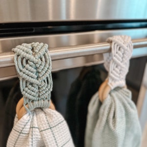 May include: Two kitchen towels hanging on a stainless steel oven handle. The towels are both light green and have a woven texture. One towel has a white macrame loop and the other has a light green macrame loop.