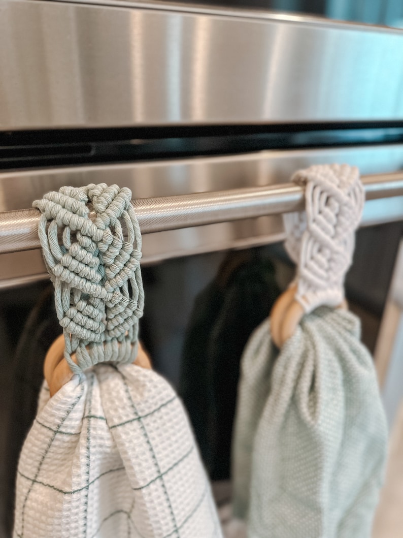 May include: Two kitchen towels hanging on a stainless steel oven handle. The towels are both light green and have a woven texture. One towel has a white macrame loop and the other has a light green macrame loop.