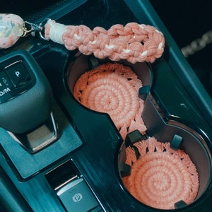 May include: Two pink crocheted cup coasters with fringe in a car's cup holder. A pink and white crocheted keychain with a floral charm hangs from the cup holder.