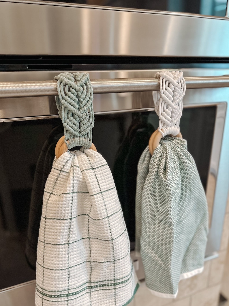 May include: Two green and white kitchen towels hanging on a silver oven rack. The towels are attached to wooden rings with macrame loops.