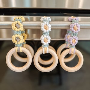 May include: Three wooden rings with crocheted flower embellishments in yellow, white, and purple. The rings are hanging from a metal bar.