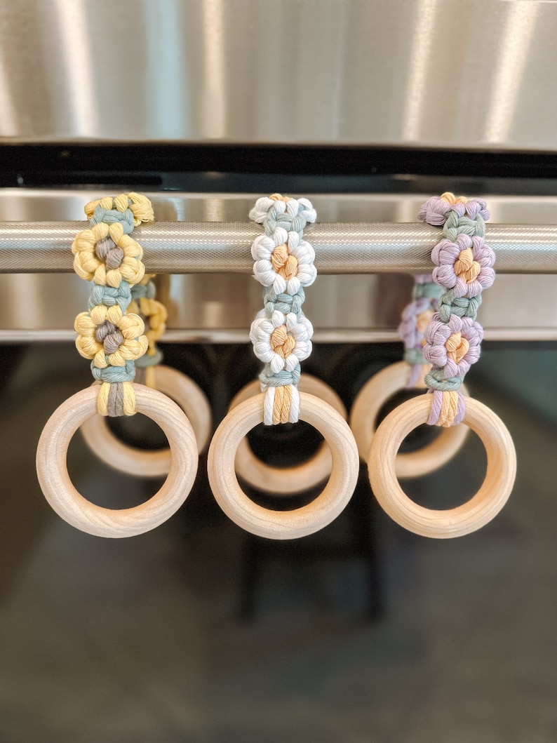 May include: Three wooden rings with crocheted flower embellishments in yellow, white, and purple. The rings are hanging from a metal bar.