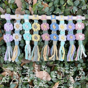 May include: Ten colorful macrame flower wall hangings with tassels. The flowers are in shades of pink, purple, blue, green, and yellow. The flowers are labeled with the names: Peony, Daisy, Orchid, Sunflower, Primrose, Iris, Daffodil, Lavender, and Rose.