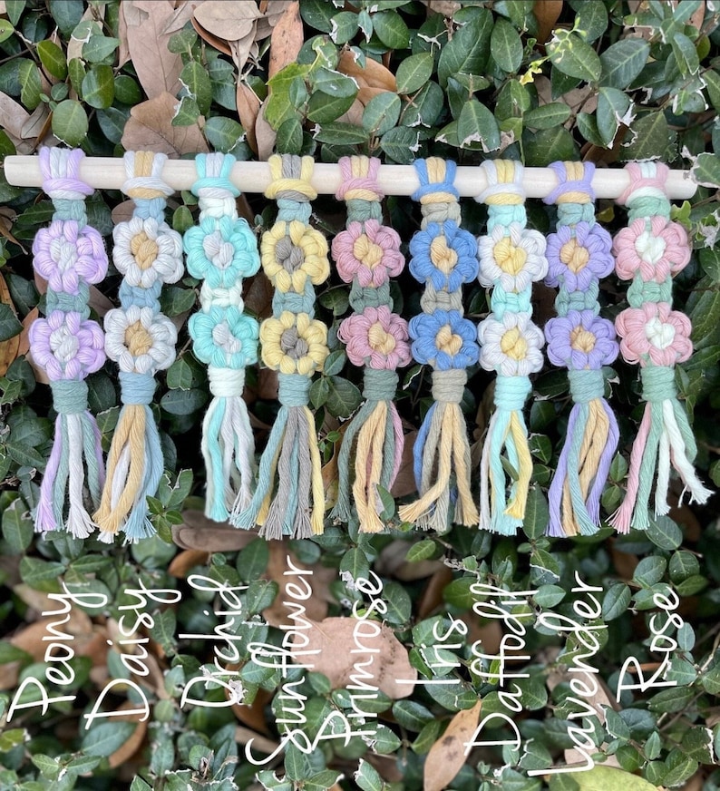 May include: Ten colorful macrame flower wall hangings with tassels. The flowers are in shades of pink, purple, blue, green, and yellow. The flowers are labeled with the names: Peony, Daisy, Orchid, Sunflower, Primrose, Iris, Daffodil, Lavender, and Rose.