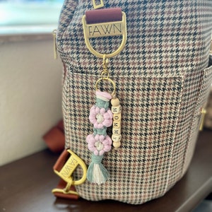 May include: A brown and white patterned diaper bag with a gold zipper and hardware. A macrame keychain with pink and green flowers and wooden beads spelling "MOMMY" hangs from the bag. A gold metal tag with the word "FAWN" is attached.