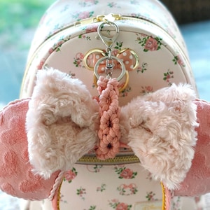 Peut inclure: Un sac à dos floral rose et blanc avec un serre-tête à nœud rose et blanc attaché à l'avant. Le serre-tête est doté d'un cordon tressé rose et blanc attaché à un porte-clés argenté avec un charme Mickey Mouse.