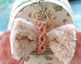 May include: A pink and white floral backpack with a pink and white bow headband attached to the front. The headband has a pink and white braided cord attached to a silver key chain with a Mickey Mouse charm.