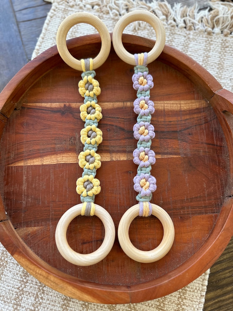 May include: Two wooden rings with macrame flower accents. The rings are natural wood and the flowers are in shades of yellow, green, and purple.