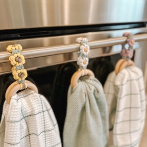 May include: Three kitchen towels hanging on a stainless steel oven handle. The towels are white with green checkered patterns, light green, and white with black checkered patterns. Each towel is hung on a wooden ring with a crocheted flower embellishment in yellow, white, and purple.
