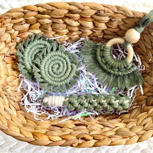 May include: A set of three green macrame baby toys in a woven basket. The set includes a teether, a rattle, and a hanging toy.