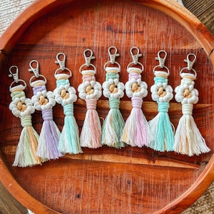 May include: Eight colorful crocheted flower keychains with tassels. The keychains are arranged in a circle on a wooden tray. The colors of the keychains are yellow, purple, green, pink, and blue.
