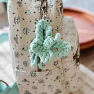 May include: A white backpack with a light blue and gold print of Disney princesses and characters. A light blue braided tassel hangs from the zipper pull.
