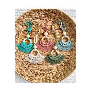 May include: Five handmade macrame car charms with wooden rings and tassels in various colors, including teal, pink, white, green, and blue. The charms are displayed on a woven wicker tray.