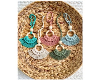 May include: Five handmade macrame car charms with wooden rings and tassels in various colors, including teal, pink, white, green, and blue. The charms are displayed on a woven wicker tray.