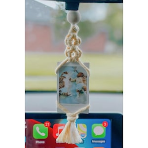 May include: A white macrame car charm with a tassel hanging from the bottom. A white photo of two cats sleeping is attached to the charm.