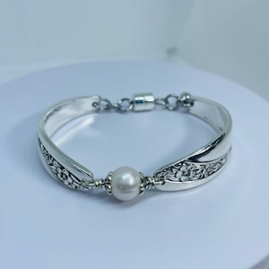 May include: A silver bracelet featuring a central white pearl and floral spoon handle design. The bracelet has a magnetic clasp and is displayed on a white surface. The spoon handles are curved and have floral patterns.