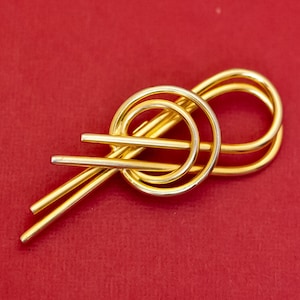 Puede incluir: Un broche abstracto dorado con un diseño anudado. El broche está hecho de metal y tiene un acabado liso y pulido.