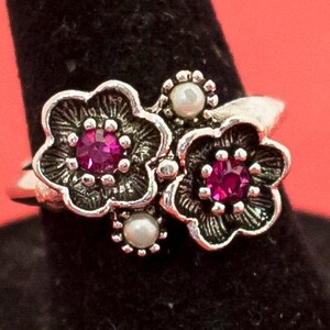 May include: A silver-toned ring featuring a floral design with two flowers and small pearl accents. Each flower has a vibrant magenta center stone. The ring is displayed against a red background.