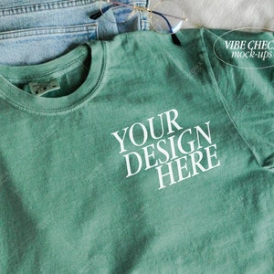 以下が含まれることがあります： 「YOUR DESIGN HERE」の白文字が入った、シーフォームグリーンのTシャツ。青いジーンズの上に置かれています。「VIBE CHECK mock-ups」の文字が右上隅にあります。金縁のメガネがTシャツの上に置かれています。