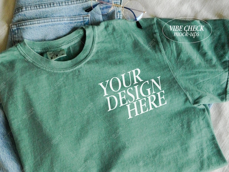 以下が含まれることがあります： 「YOUR DESIGN HERE」の白文字が入った、シーフォームグリーンのTシャツ。青いジーンズの上に置かれています。「VIBE CHECK mock-ups」の文字が右上隅にあります。金縁のメガネがTシャツの上に置かれています。
