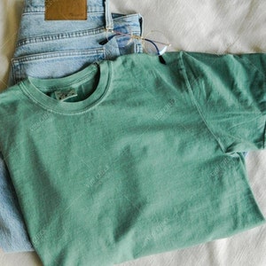 以下が含まれることがあります： ミントグリーンのTシャツ、ライトウォッシュデニムのジーンズ、丸いフレームの眼鏡を組み合わせたカジュアルなアンサンブル。白いテクスチャのある表面に配置され、リラックスした日常的な美学を作り出しています。