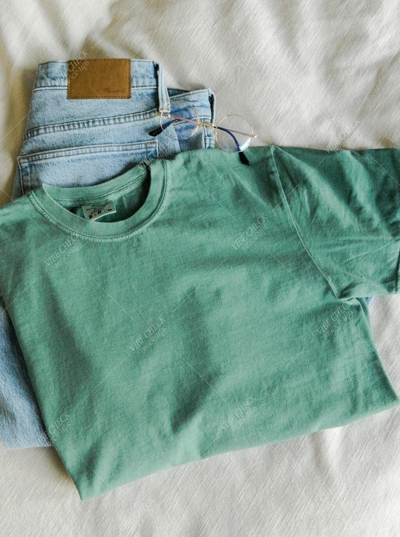以下が含まれることがあります： ミントグリーンのTシャツ、ライトウォッシュデニムのジーンズ、丸いフレームの眼鏡を組み合わせたカジュアルなアンサンブル。白いテクスチャのある表面に配置され、リラックスした日常的な美学を作り出しています。