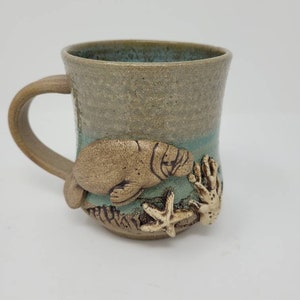Peut inclure: Une tasse en céramique avec un motif de lamantin brun, une étoile de mer et du corail. La tasse est émaillée en brun clair et bleu.