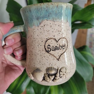 Pode incluir: Uma caneca de cerâmica branca com um esmalte salpicado e um design em forma de coração que diz "Sanibel". A caneca tem um esmalte azul claro na parte superior e apresenta um design de conchas na parte inferior.