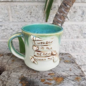 Könnte beinhalten: Eine weiße Keramiktasse mit einer türkisfarbenen Glasur innen und einer hellgrünen Glasur außen. Die Tasse hat einen Henkel und sagt "Ich frage mich, ob der Strand mich vermisst?" mit Muscheln und einem Seestern-Design.