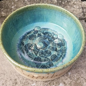Peut inclure: Un grand bol rond en céramique avec un glaçage turquoise et un motif de coquillages noirs. Le bol a un bord brun clair et le mot "Camille" est écrit au fond.