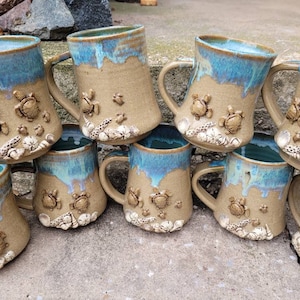 May include: A stack of eight ceramic mugs with a light brown glaze and blue accents. Each mug features a sea turtle design with seashells and starfish.