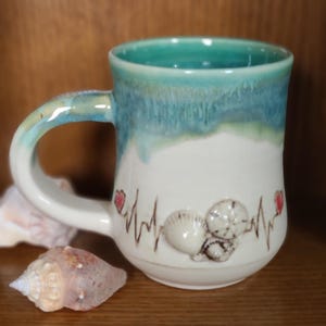 Può includere: Una tazza in ceramica bianca con una glassa turchese e un design a conchiglia. La tazza ha una linea di battito cardiaco con due cuori rossi e un design a conchiglia.