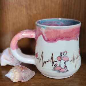 Può includere: Tazza in ceramica bianca con una glassa rosa e blu. La tazza ha un cuore e una linea di fenicotteri rosa con una linea di battito cardiaco sottostante. La tazza è appoggiata su una superficie di legno.