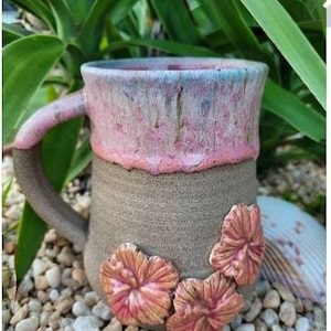 Può includere: Una tazza in ceramica con una glassa rosa chiaro e blu e decorazioni floreali tridimensionali. La tazza ha un manico marrone e si trova su un letto di piccole pietre bianche.