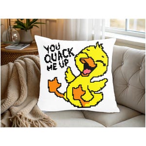 Może przedstawiać: Biała kwadratowa poduszka z pikselową grafiką żółtej kaczki. Kaczka ma otwarty dziób i pomarańczowe stopy. Tekst "YOU QUACK ME UP" znajduje się nad kaczką w czarnych pikselowych literach. Poduszka leży na beżowej sofie.