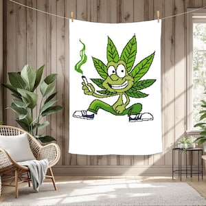 Op de afbeelding: Een witte rechthoekige wandtapijt met een cartoon cannabisblad karakter. Het groene karakter houdt een brandende sigaret vast en blaast groene rook uit. Het draagt een groene broek en witte schoenen met blauwe details. Het tapijt hangt aan een waslijn.