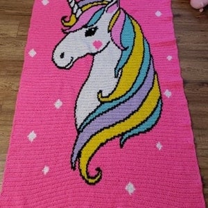 May include: A pink crocheted blanket featuring a white unicorn design. The unicorn has a rainbow mane and tail.