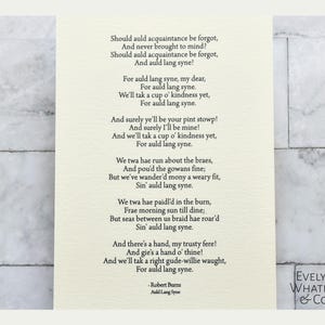 May include: A vintage-style print of a poem titled "Auld Lang Syne" by Robert Burns. The poem is printed in black ink on a white background. The poem is about the importance of remembering old friends and the passage of time. The print is framed with a black border and includes the name "Evelyn Whatley & Co."