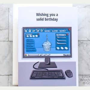 May include: A birthday card with the text "Wishing you a solid birthday." The card features a cartoon illustration of a computer monitor displaying a cupcake with a lit candle. A keyboard and mouse are below the monitor.