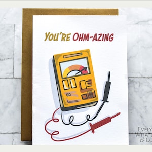 May include: A white card with the text "You're Ohm-azing" in red and black lettering. A yellow and black cartoon illustration of a multimeter with red and black wires is below the text. The card has a brown envelope behind it and the text "Evelyn Whatley & Co." in black lettering in the bottom right corner.