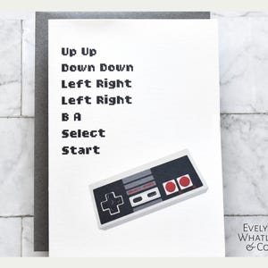 May include: A white greeting card with a gray border features the text "Up Up Down Down Left Right Left Right B A Select Start" in a pixelated font. A black, gray, and red video game controller is at the bottom. The Evelyn Whatley & Co. logo is in the bottom right corner.