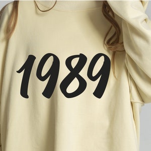 May include: A cream colored sweatshirt with the number 1989 printed in black on the front.