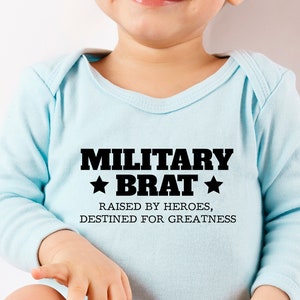 Può includere: Un body per neonato di colore azzurro chiaro con testo nero che recita "Military Brat" con una stella su entrambi i lati della parola "Brat". Il testo sottostante recita "Raised by heroes, destined for greatness."