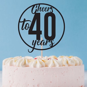 Op de afbeelding: Een zwarte taarttopper met de tekst "Cheers to 40 years" in een cirkel. De topper zit bovenop een roze geglazuurde taart.