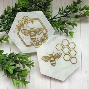 Può includere: Due sottobicchieri in marmo bianco con disegni di api e favi dorati. L'ape sul sottobicchiere a sinistra è circondata da fiori. Il disegno a nido d'ape sul sottobicchiere a destra ha la forma di un esagono.