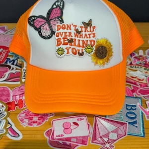 Peut inclure: Casquette de camionneur orange et blanche avec un papillon et un patch de tournesol. La casquette porte l'inscription "DON'T TRIP OVER WHAT'S BEHIND YOU". Elle a un dos en filet et une visière incurvée.