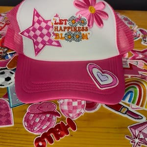 May include: A pink and white trucker hat with a pink mesh back. The front features a star and flower patch, along with the phrase "LET HAPPINESS BLOOM". Additional patches with hearts, flowers, and the word "HELLO" are scattered around.