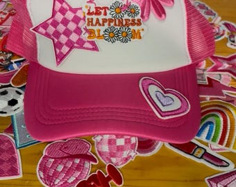 May include: A pink and white trucker hat with a pink mesh back. The front features a star and flower patch, along with the phrase "LET HAPPINESS BLOOM". Additional patches with hearts, flowers, and the word "HELLO" are scattered around.