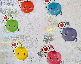 May include: Six colourful apple stickers with cartoon designs. Each apple is a different colour: red, blue, green, purple, yellow, and pink. They have smiling faces, small arms, and a speech bubble with a heart. The stickers are on a silver, iridescent background.